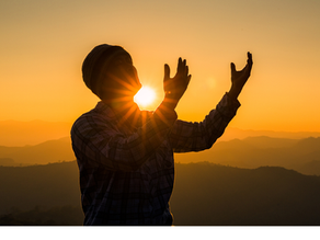 Es un hombre levantando las manos al cielo y de fondo se ve la puesta del sol de color naranja.