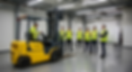 Bir grup öğrenci veya yeni çalışan, forklift eğitim kursunda sarı renkli bir forkliftin nasıl kullanılacağını öğreniyor. Eğitim alanı geniş ve aydınlık olup, duvarlarda forkliftin çalışma mekanizmasını gösteren teknik çizimler bulunuyor. Eğitmen, forkliftin kontrollerini açıklarken öğrenciler dikkatle dinliyor. Tüm katılımcılar, iş güvenliği yönetmeliklerine uygun olarak reflektörlü sarı yelekler giymiş. Forkliftin biraz yıpranmış olması, eğitim amaçlı düzenli olarak kullanıldığını gösteriyor. Bu eğitim programı, forklift operatörlerinin güvenli ve verimli bir şekilde çalışmasını sağlamak için tasarlanmış.