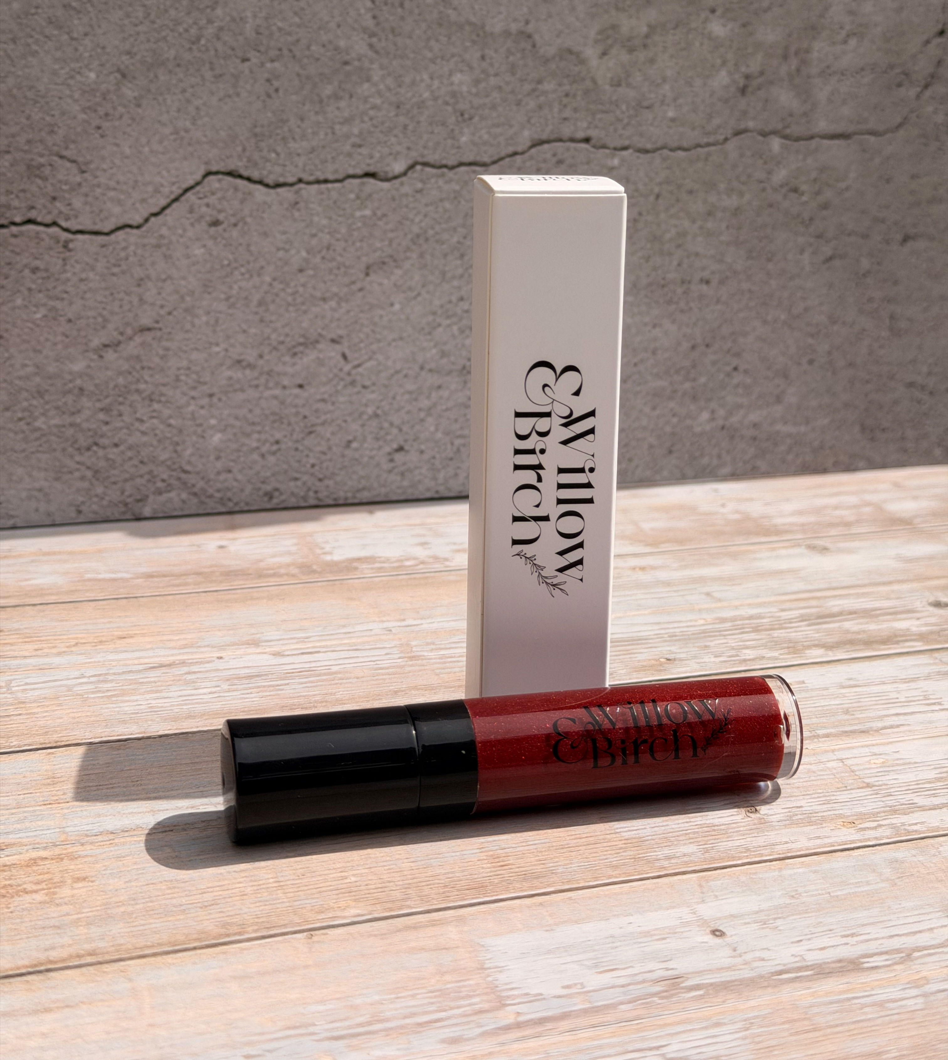 A tube of red coloured lip gloss lying down on light wooden surface with white lip gloss box standing next to it.