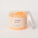 An open clear plastic pot on a white background showing the orange scrub contents with orange coloured label on the front. The silver lid is stood on its edge leaning onto the jar.
