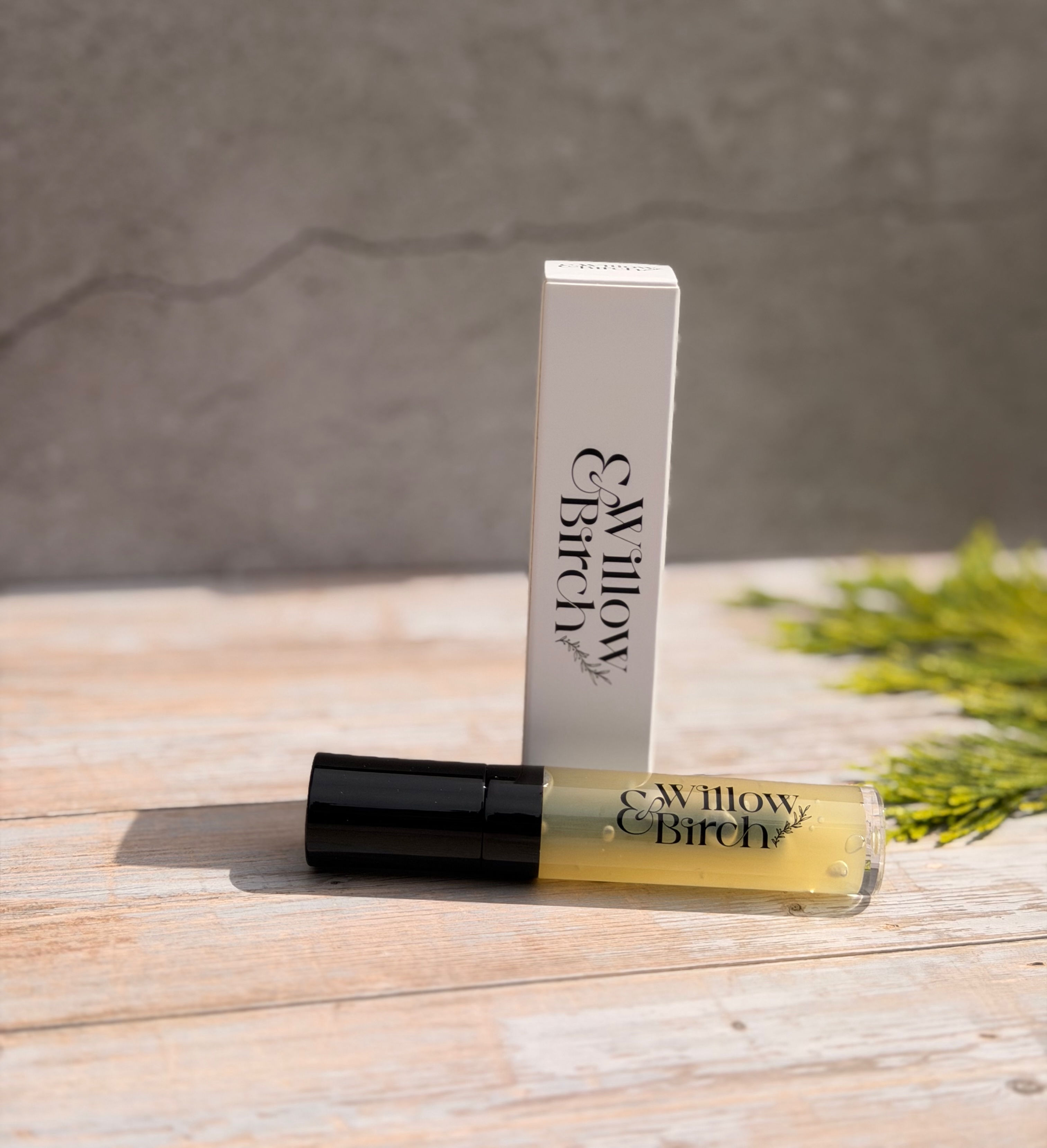 A tube of cream coloured lip gloss lying on its side in front of a white lip gloss box on a light wooden surface