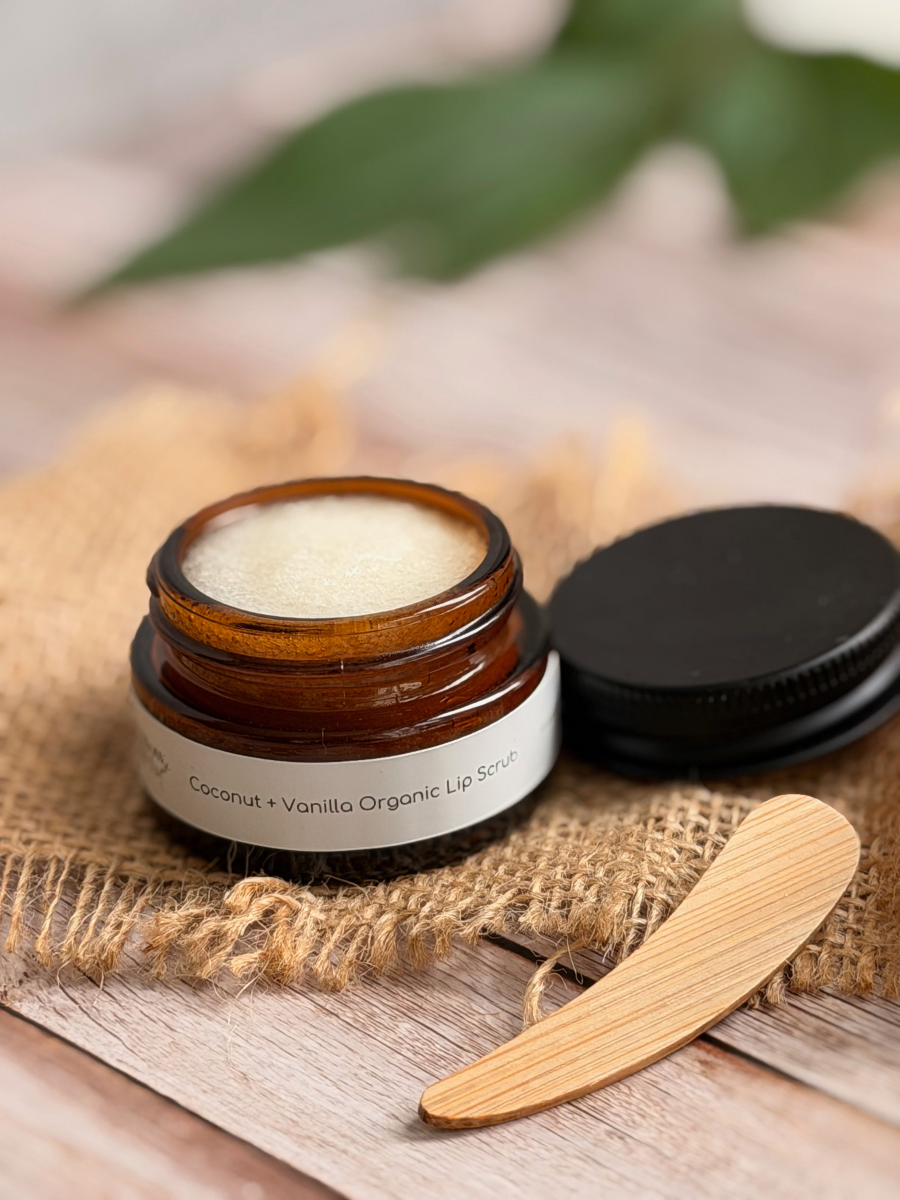 A small brown glass pot open showing lip scrub inside, black lid lying beside it along with small wooden spatula
