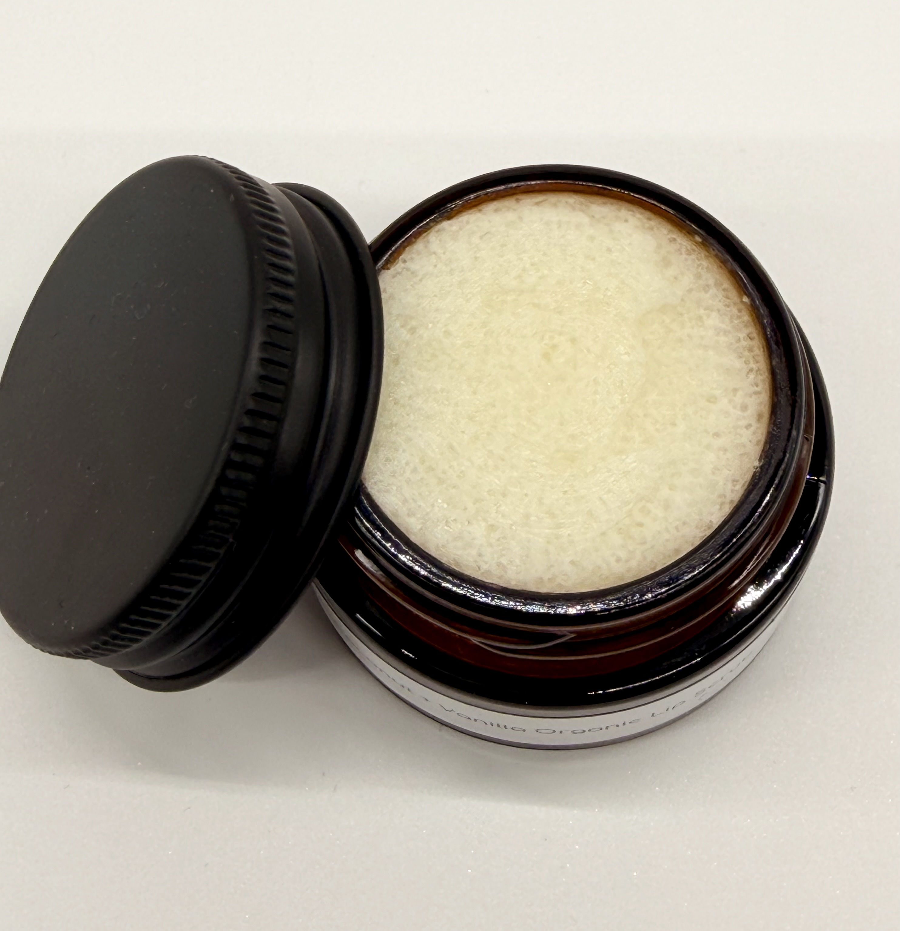 A small round brown glass jar with cream coloured lip scrub contents. Lid is lying to the side of the pot
