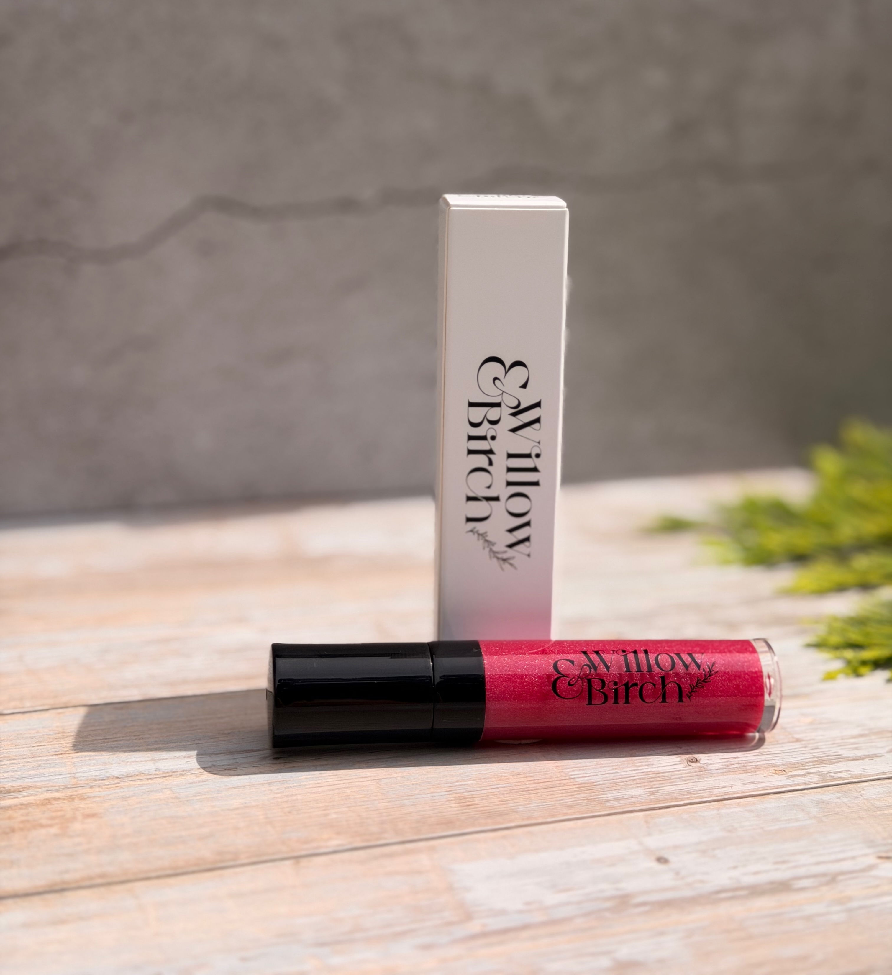 A tube of pink coloured lip gloss lying on light wood surface with white lip gloss box standing behind it.