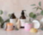 A bottle of bath foam in brown glass bottle with silver lid, a bottle of shower gel in brown glass bottle with black plastic pump top. Two plastic pots falloff whipped soap one oink and one orange along with a silver aluminium pot and the flower shaped lotion bar lying in front of it. Stood on a pink coloured background with eucalyptus branches.