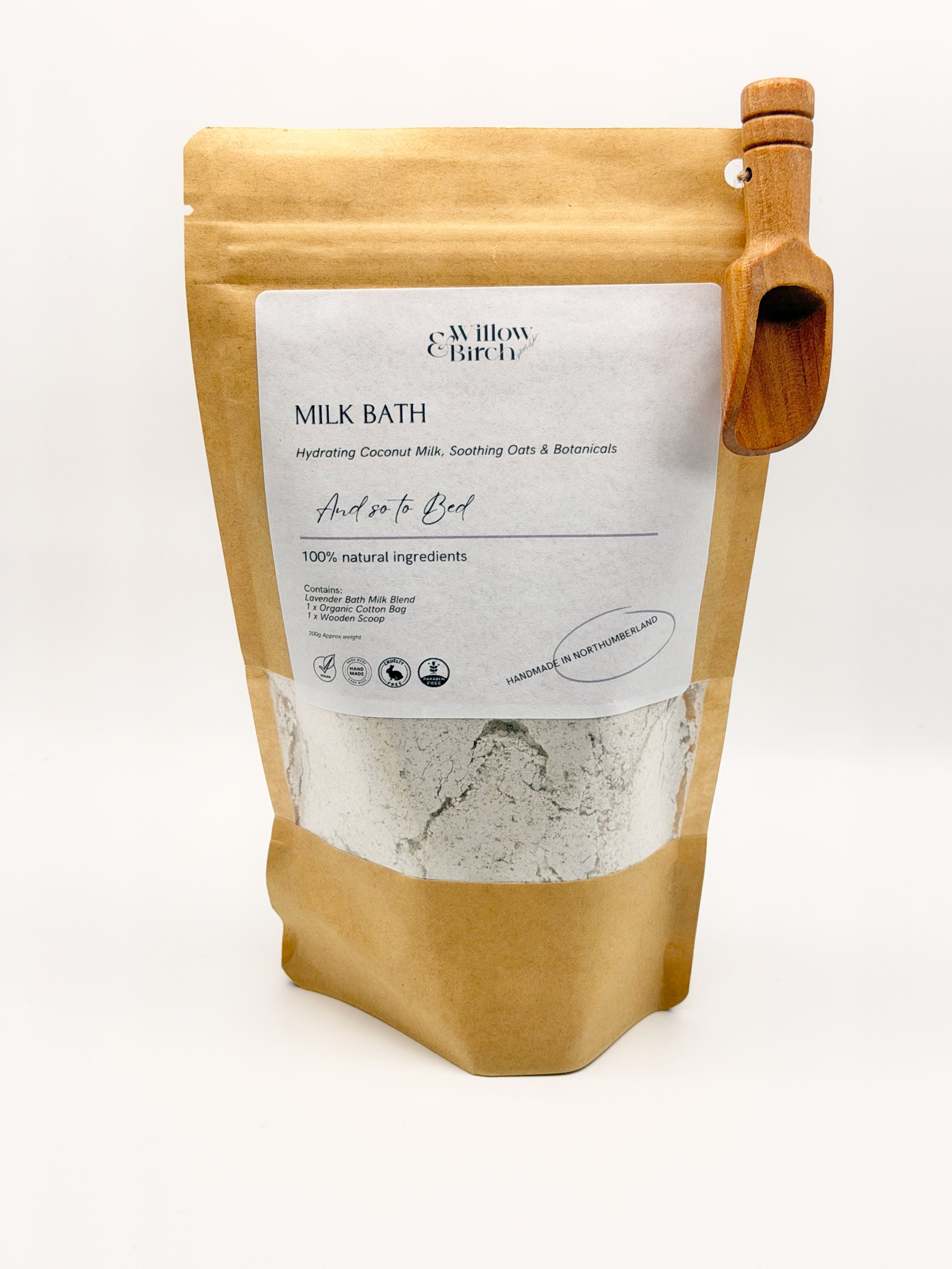 Brown bag with clear window, with bath milk inside a small wooden scoop is attached to the corner of the bag