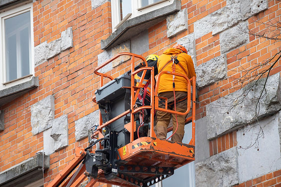 Two construction workers in safety gear stand on an orange lift, working on a brick building facade
