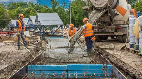 Construction workers in safety vests and helmets are pouring concrete from a mixer truck into a road framework