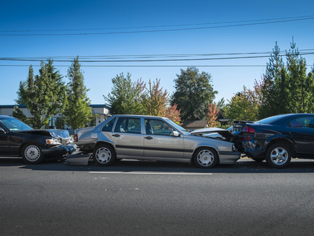 Three-car rear-end collision on a street. The silver car in the middle has front and rear damage, while the black cars on either side show impact damage to their bumpers.