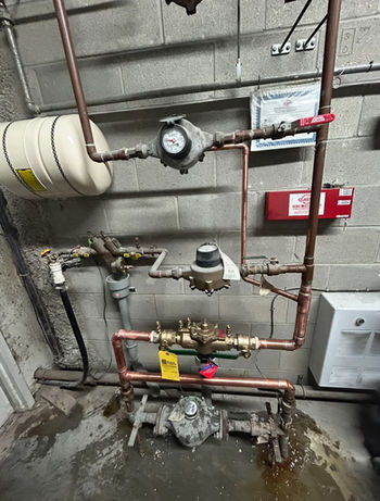 A commercial plumbing setup featuring copper piping, a large expansion tank, two water meters, and a backflow prevention assembly mounted on a gray cinder block wall.