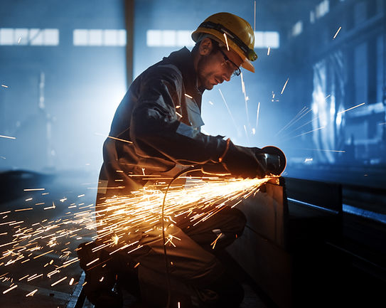 A worker in protective gear and a yellow helmet grinds metal, creating a shower of bright sparks in a dimly lit industrial setting
