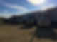 Five large trucks, including dump trucks, are parked in a row on a dusty lot beside a warehouse under a bright