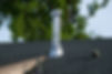 Shiny metal chimney vent on a shingled roof, with tall green trees in the background under a clear blue sky
