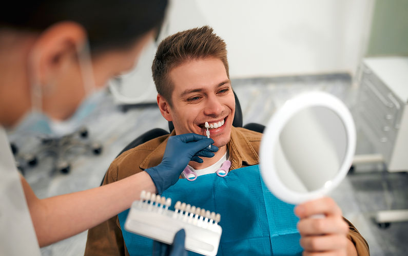 A smiling man in a dental office holds a mirror, while a dentist uses a shade guide to match his teeth color