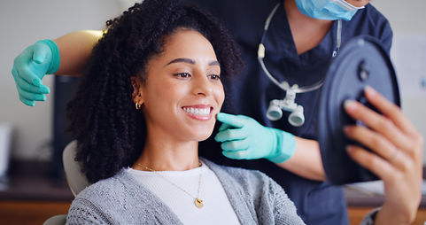 A woman with curly hair smiles while holding a mirror, sitting in a dental chair