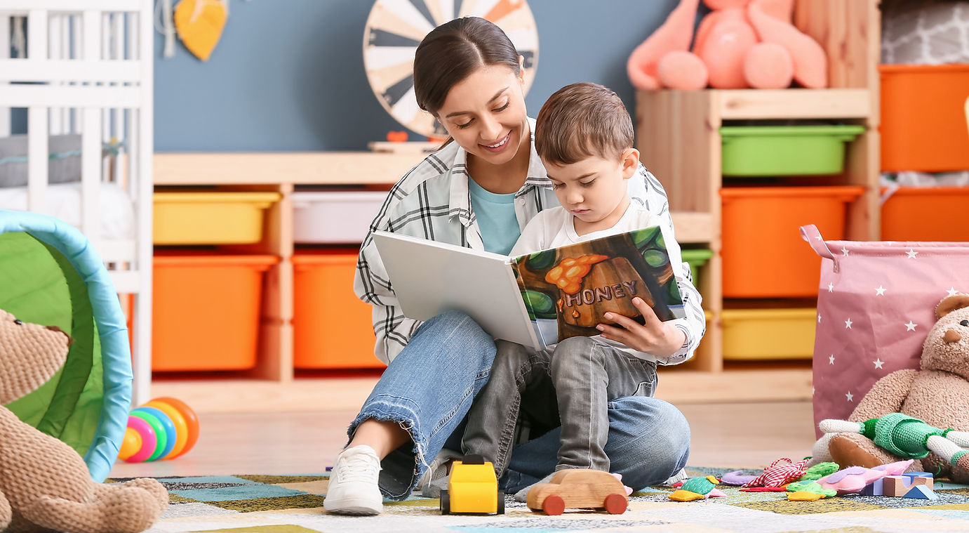A woman and child sit on the floor in a playroom reading a book together