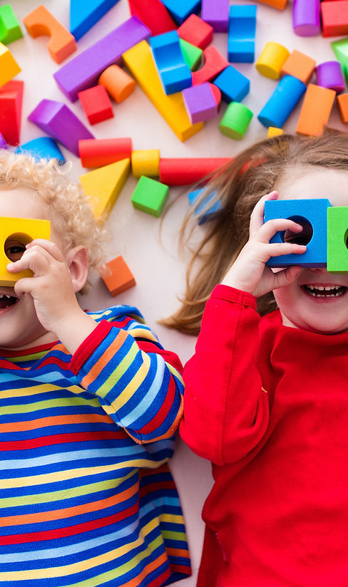 Two children lying on the floor smile while holding colorful toy blocks to their eyes
