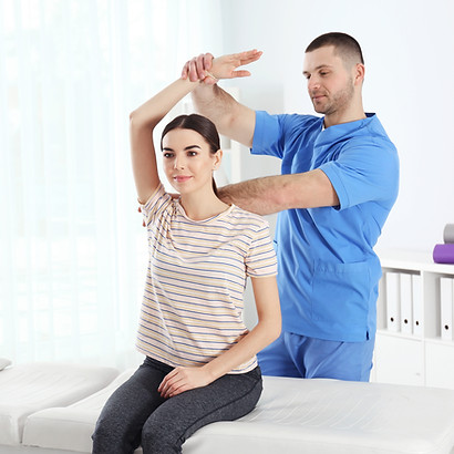 A physical therapist in blue scrubs assists a woman in a striped shirt with arm exercises