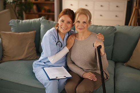 A smiling nurse in blue scrubs sits beside an elderly woman