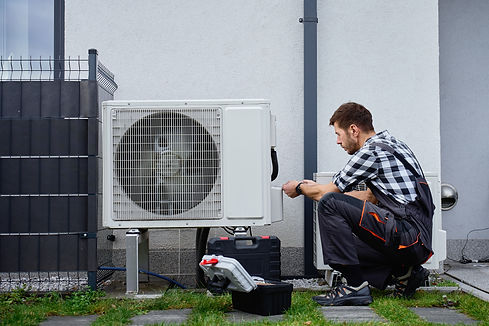 A technician in checkered shirt and overalls kneels beside an outdoor air conditioning unit, working with tools