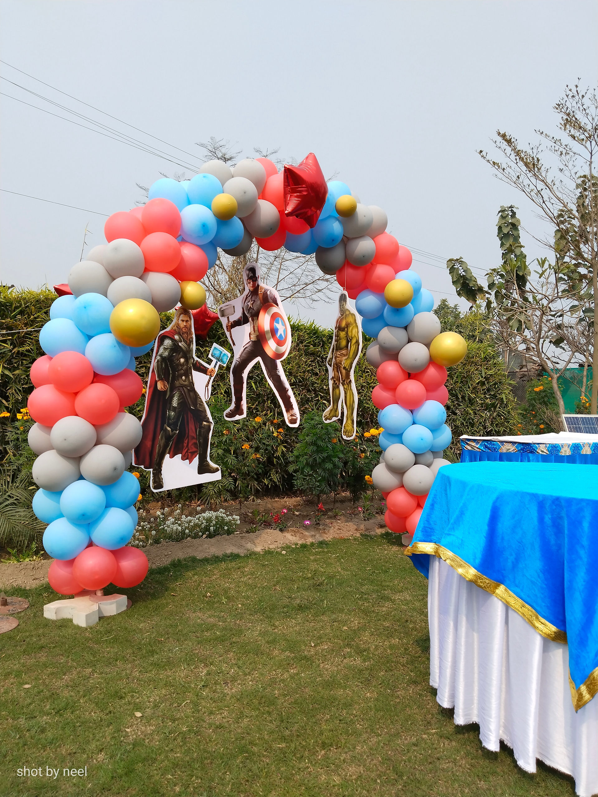 Avengers theme balloon arch on a lawn with superhero cutouts in red, sky blue, grey and gold by Birthday Bazaar