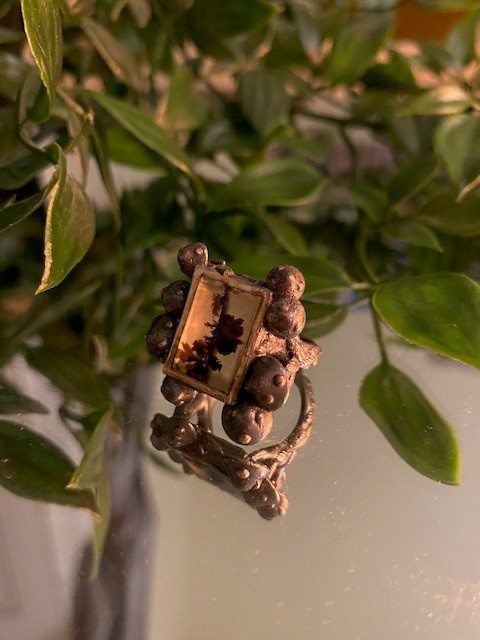 A bronze ring in the shape of a clump of mushrooms set on a mirrored surface with leaves in the background.