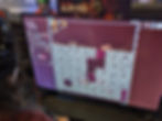 A photograph of Nate, a brown-haired person playing at an Everdeep Aurora demo station. The game is Everdeep Aurora, as signified by a small banner image at the top of the screen that shows an adorable black cat diving downward with a drill pointed at the ground. In the game, a small cat can be seen drilling into cubes in the ground.