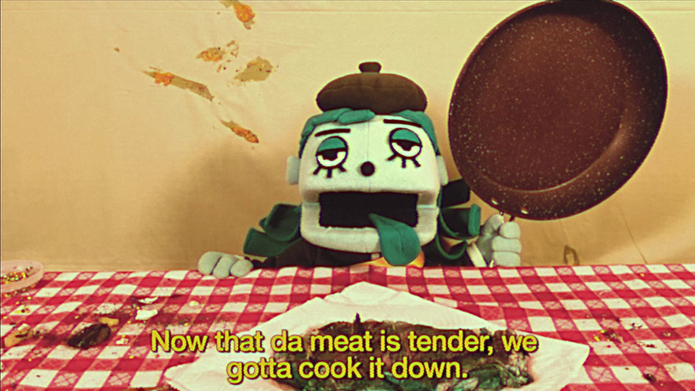 An in-game screenshot of Great God Grove. It depicts one of its live-action cutscenes featuring a puppet character from the Bizzyboys gang. A lazy-eyed green tinted puppet with its tongue out and wearing a beret, wields a frying pan. In front of them, a strange blue-tinted steak can be seen on a plate. Subtitles show they're saying "Now that da meat is tender, we gotta cook it down."