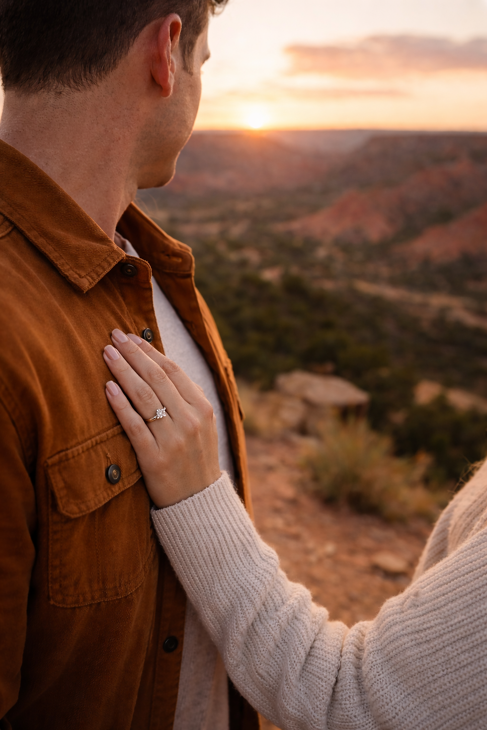 Man in jacket with woman's hand on his chest featuring an engagement ring. Scenic sunset view over Palo Duro canyon State park in the background. Romantic mood, recently engaged in a Palo Duro Canyon Valentines day proposal.
