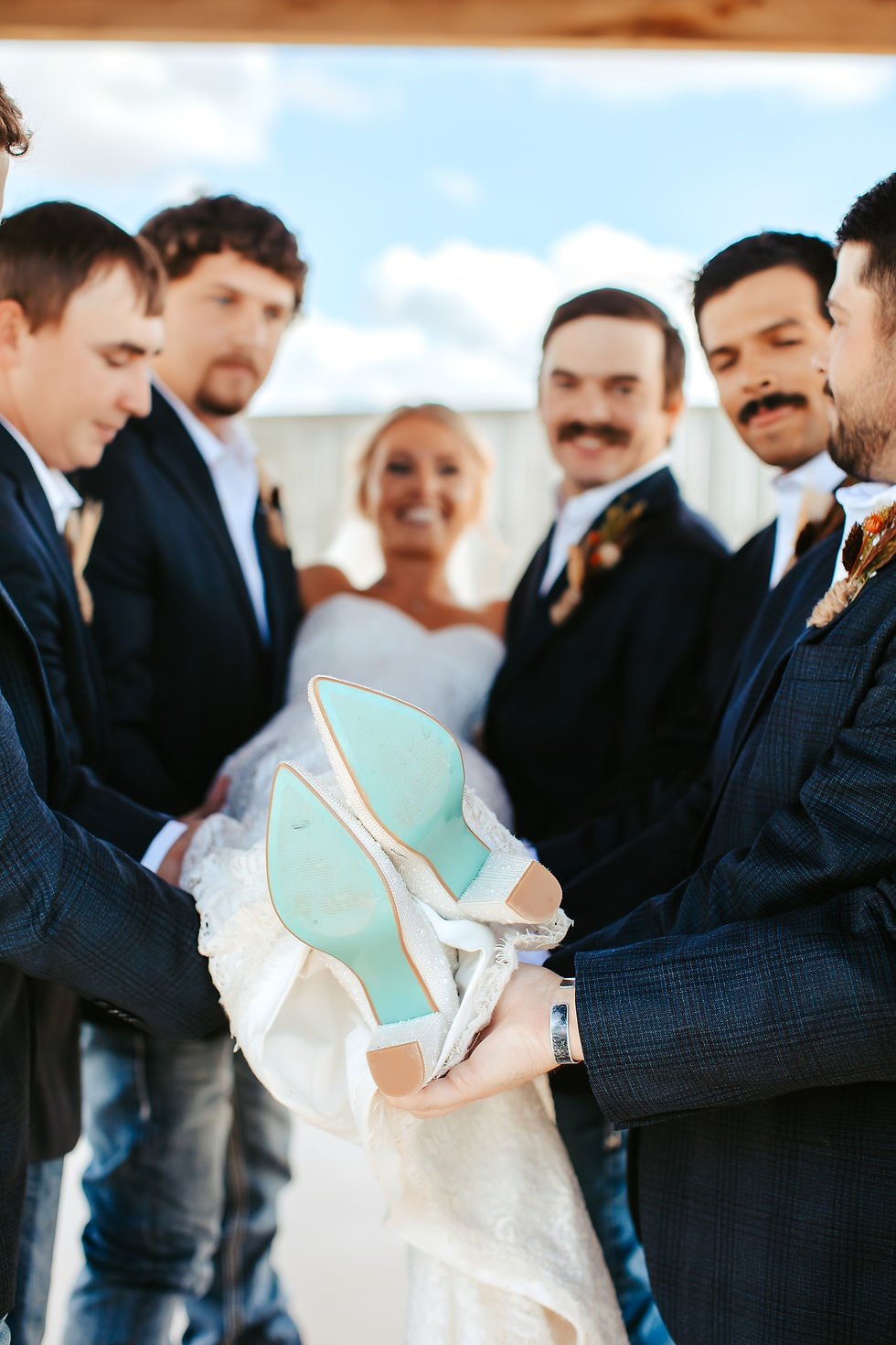 A bride in a white dress is playfully lifted by five groomsmen in suits. Sky background, turquoise shoe soles visible, joyful atmosphere at wedding venue near amarillo, rustic barn venue near amarillo for fall wedding ceremony