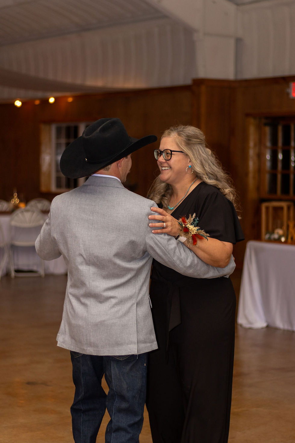 Groom in cowboy hat and gray jacket dances with a smiling woman in black. Wooden interior, joyful mood, white chairs in the background at amarillo wedding venue