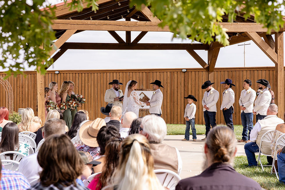 Bride and groom stand under a wooden pavilion at cornerstone ranch event center in Amarillo texas exchanging vows, with bridesmaids and groomsmen in hats, amid seated guests and greenery. Fall wedding outdoors in amarillo