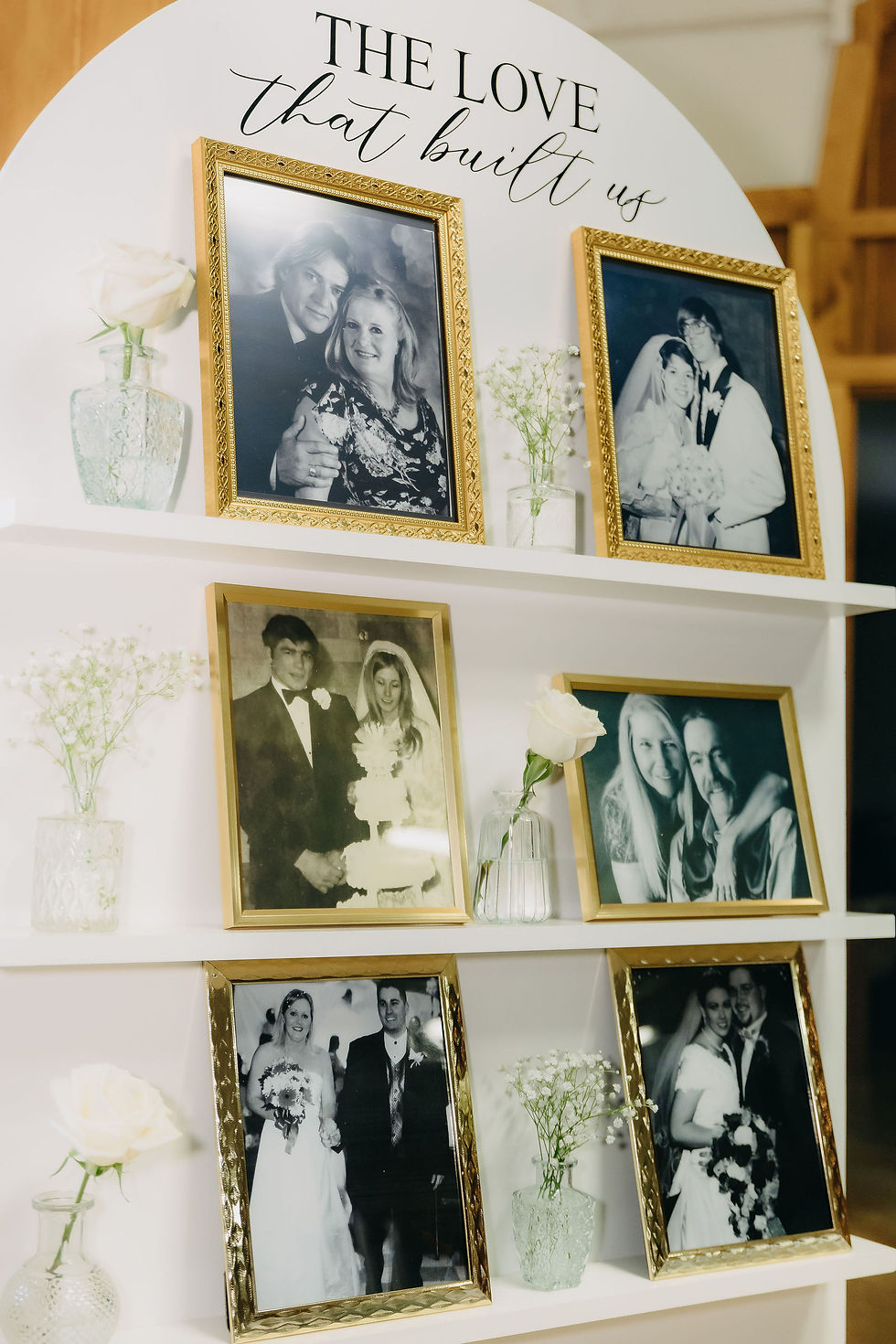 Framed black-and-white wedding photos on shelves with glass vases of white roses. Text above reads: The Love That Built Us. Beautifully decorated Texas wedding venue