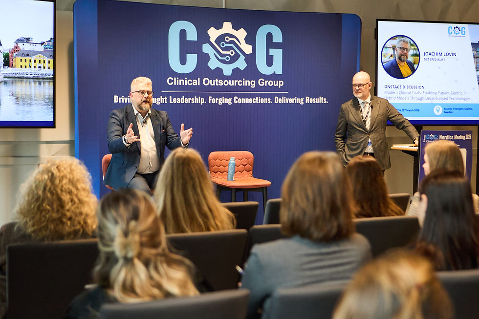 Speaker gestures on stage at Clinical Outsourcing Group event. Audience listens, blue backdrop with logo and text, second speaker stands.
