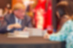 Man in a suit talking to a woman across a table with stacked books. Background shows red decoration. Glass of drink nearby.