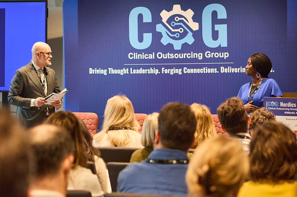 Man and woman speaking at a conference. Audience listens in a room with a blue backdrop showing "Clinical Outsourcing Group" and tagline.