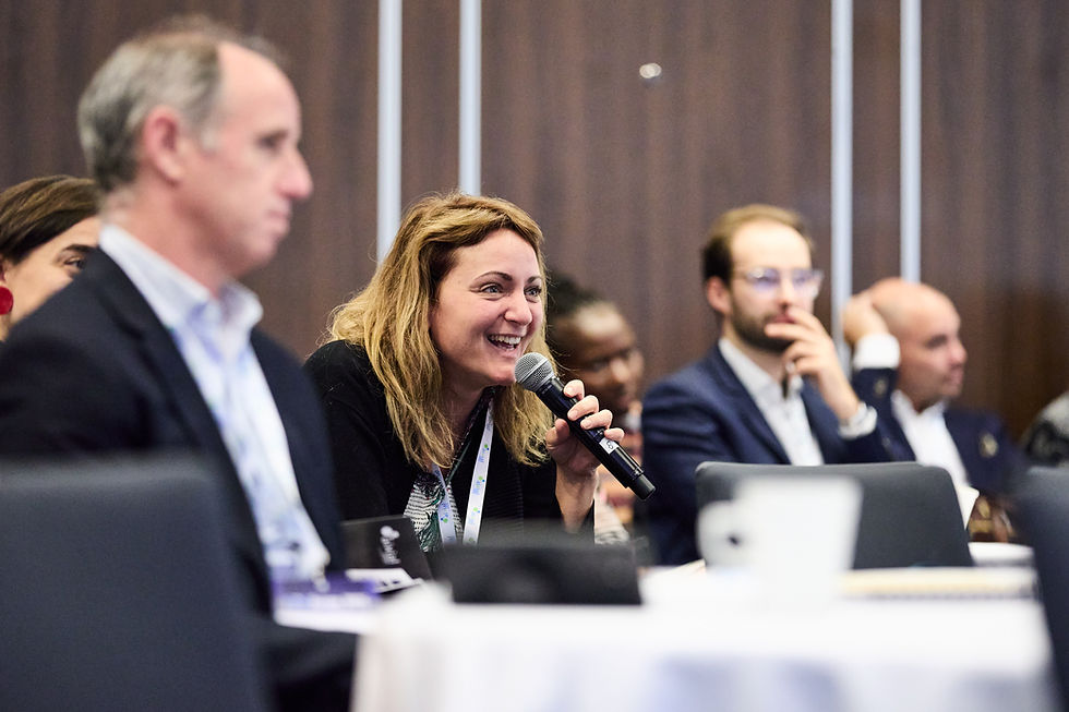 Woman with a microphone smiles during a conference panel discussion. Blurred attendees in business attire sit behind her. Warm, professional setting.