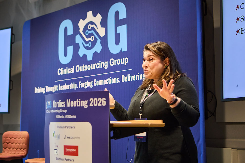 A woman speaks at a podium during the Nordics Meeting 2026. Blue backdrop with "Clinical Outsourcing Group" logo. Presentation screen nearby.