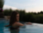 Woman in a pool, eyes closed, at sunset. Lush greenery surrounds the water. Calm and serene atmosphere.