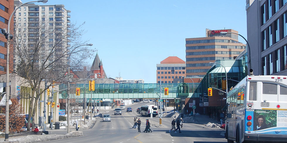 Urban street scene with people crossing at a traffic light. A bus is turning, and high-rise buildings and a glass skywalk are visible.
