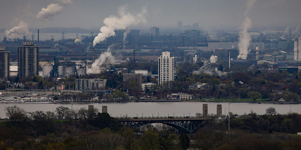 City skyline with factories emitting smoke, a river in the foreground, and a bridge surrounded by trees. Overcast skies set a gloomy mood.