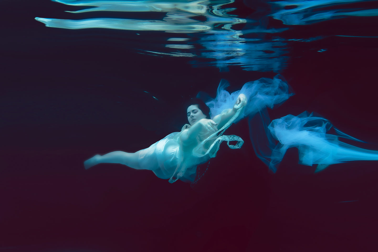 A woman in a nightgown with a long string of pearls floats through a dark blue abyss underwater. Fine art underwater photographer, Bucks County, PA, Lexi Bird Photography