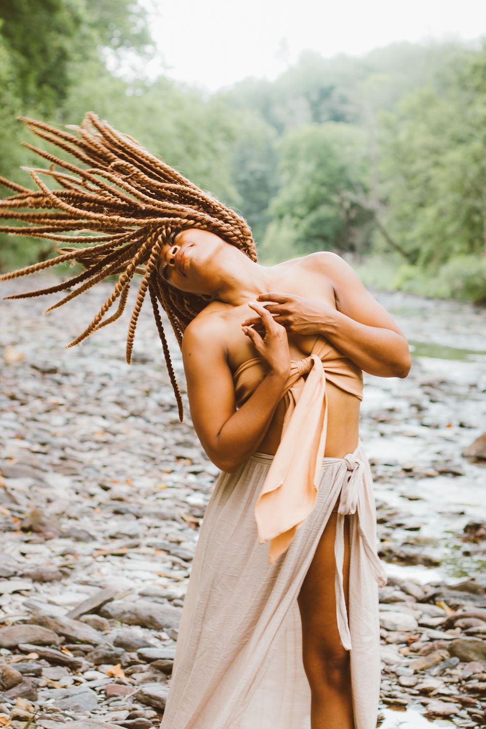 A woman with long dreadlocks and earthy clothes tosses her hair while standing in front of a rocky creek. Emotional fine art portraits in nature, New Hope, Bucks County, PA, Lexi Bird Photography