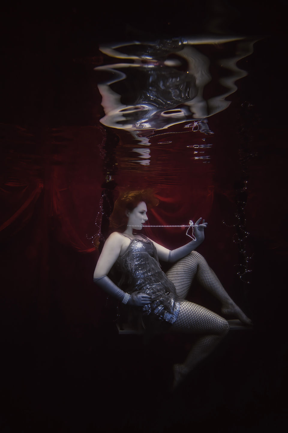 A woman dressed like a flapper or a vintage showgirl sits on a swing holding a string of pearls as she floats underwater. Underwater portraits, Greater Philadelphia, PA, Lexi Bird Photography