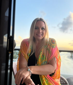 A photo of a blonde women facing the camera smiling. Wearing a bright multicoloured top with black underneath. Background is a view of the Nerang river from the top floor.