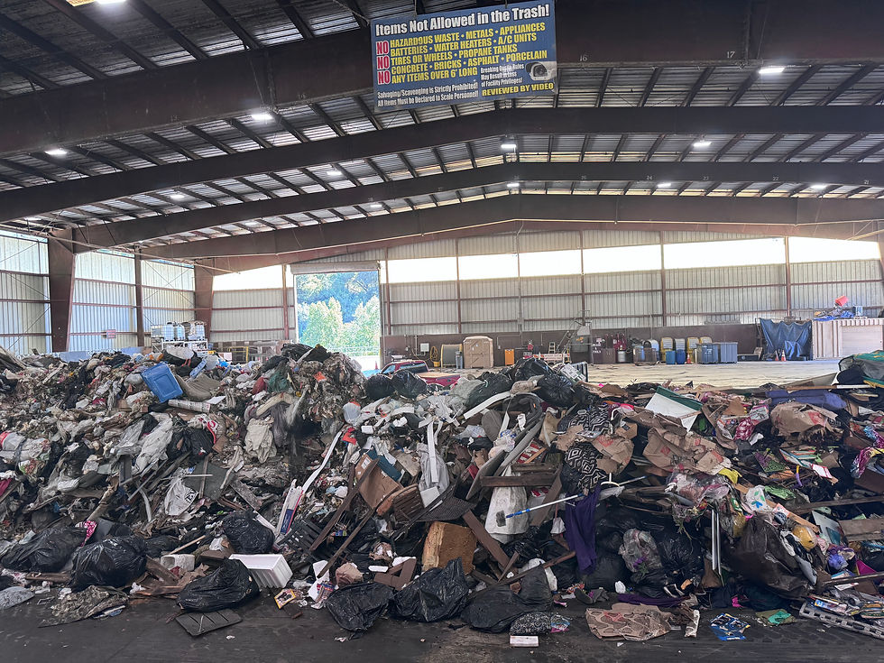 Piles of assorted trash in a large warehouse. Overhead sign lists items not allowed in trash. Sunlight enters through open door, revealing trees.