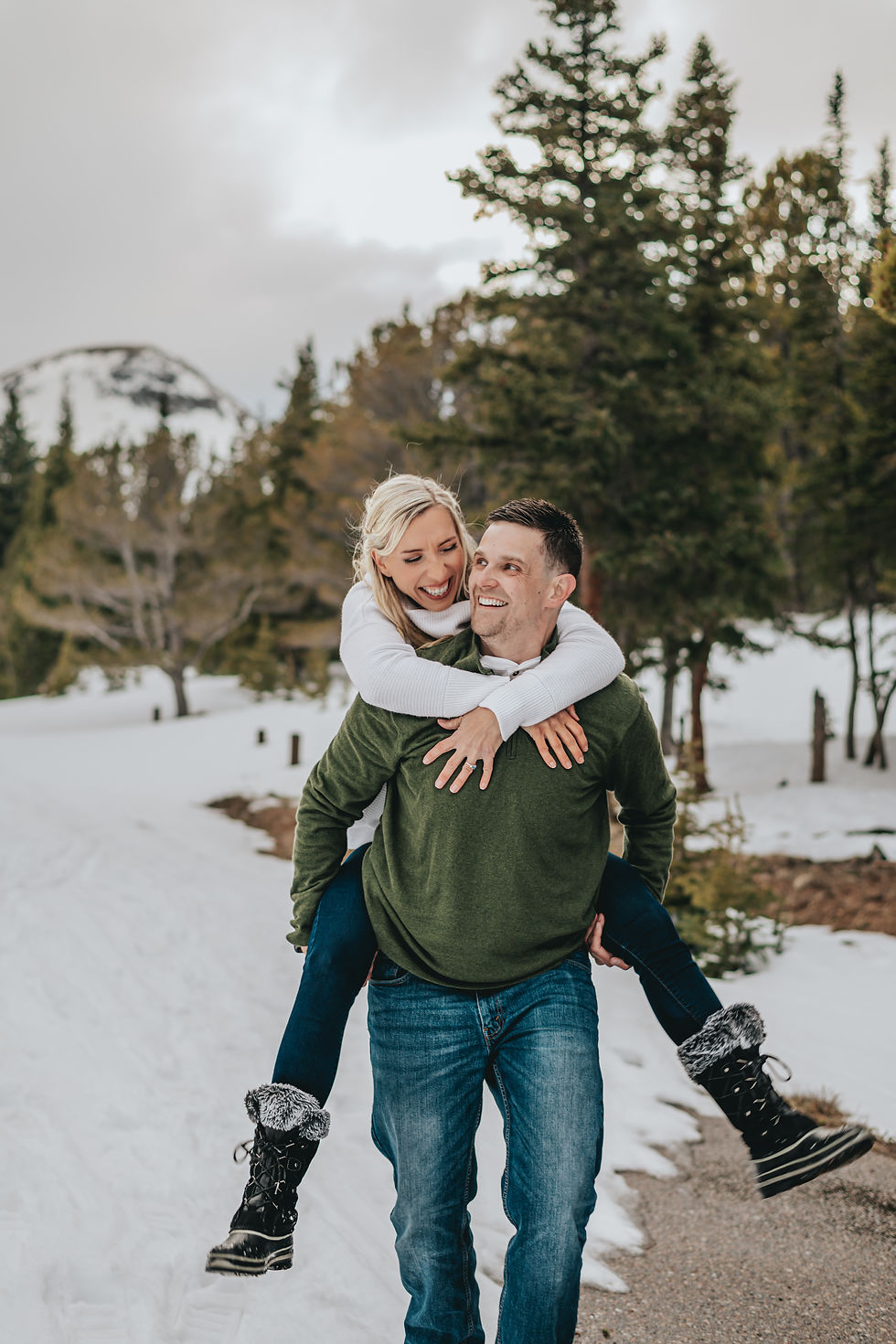 happy-engaged-couple-woman-riding-mans-back-snowy-mountains-pine-trees