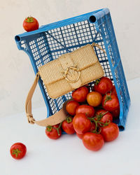 Natural straw summer bag with gold details and beige leather strap, placed on an overturned blue plastic crate surrounded by ripe red tomatoes on a white background