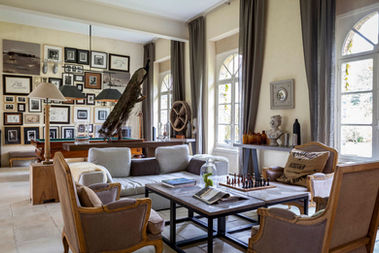 Très grand salon avec au premier plan une table à jouer avec trois fauteuils et un canapé. Au fond il y a un billard et des cadres accroché au mur.Trois fenêtres éclaire la pièce