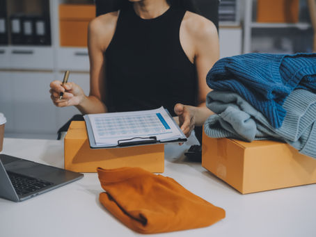 Person in black top checks a clipboard with financial information beside folded sweaters on boxes in a clothing office, with a laptop and coffee.